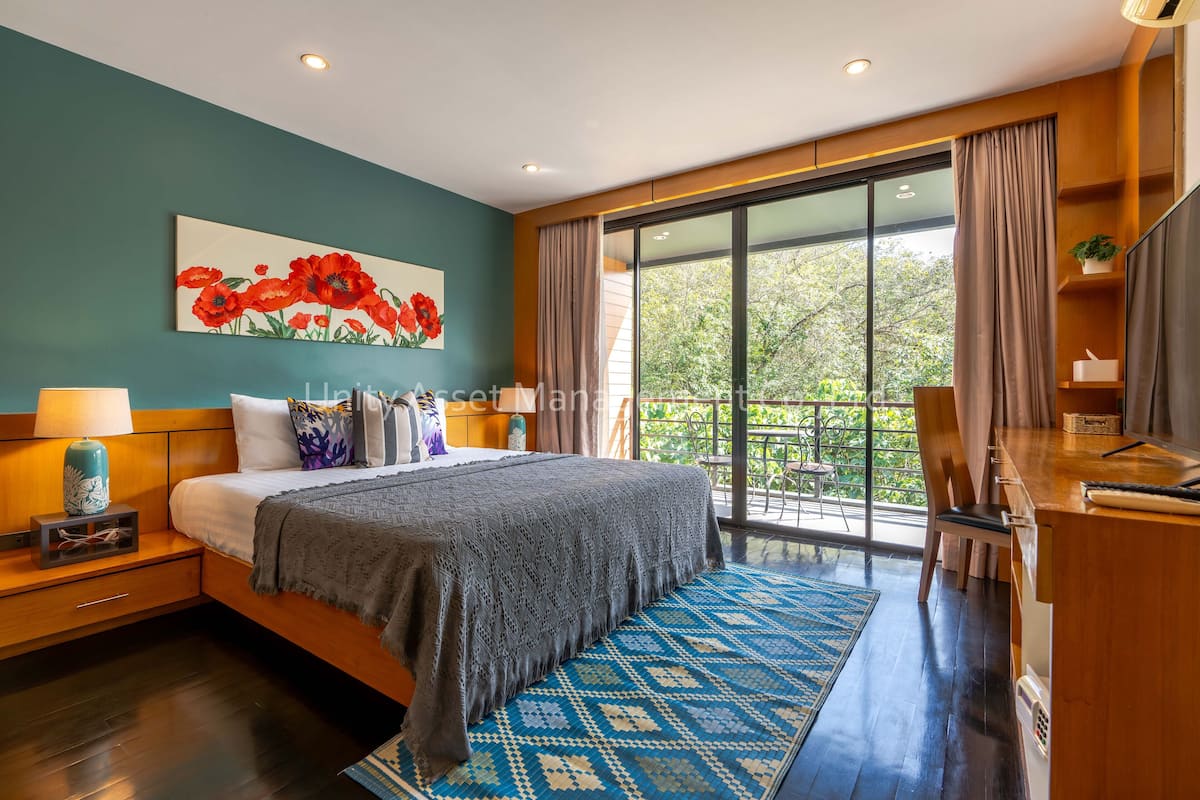 A modern bedroom with a large bed, a view of greenery through the window, and a wooden desk with a chair.