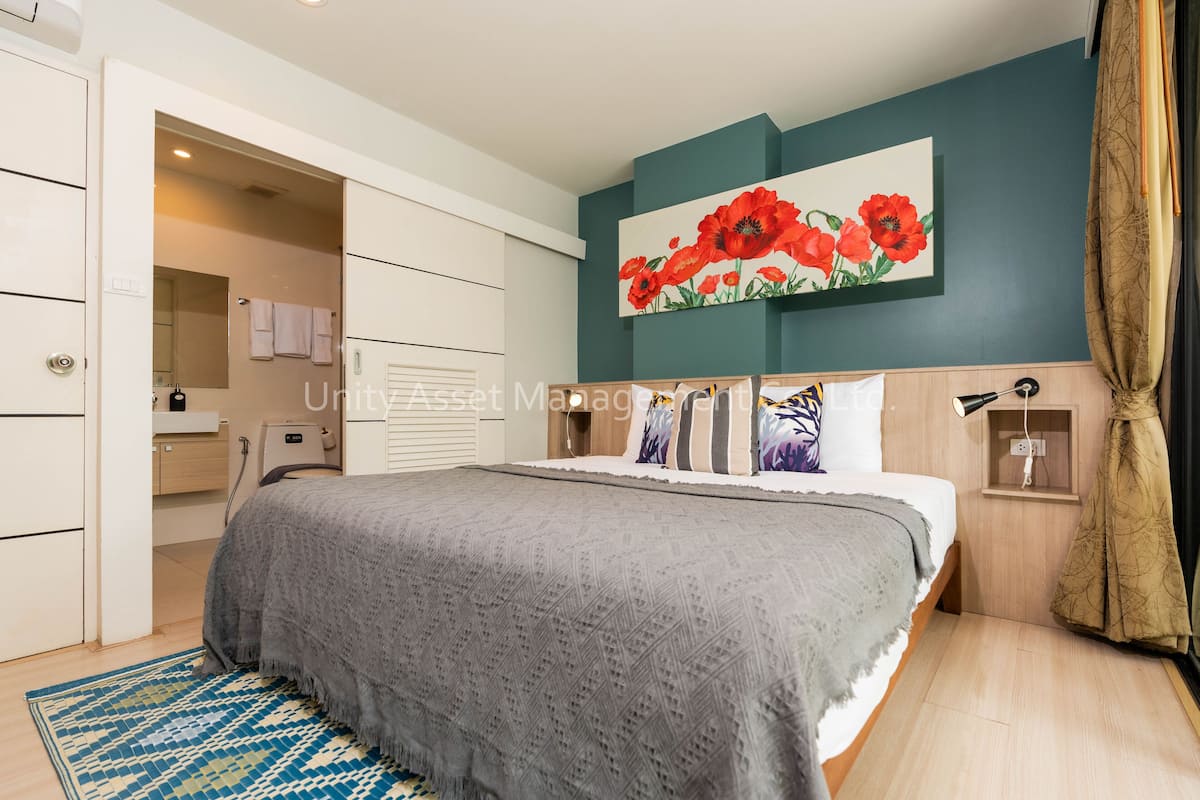 A bedroom with a bed, a nightstand, a wall-mounted lamp, a painting of red flowers, and a door leading to a bathroom.