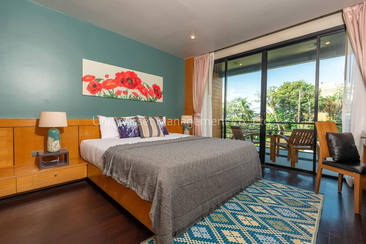 A bedroom with a large bed, a painting of red flowers, a view of greenery through a sliding door, and a patterned rug.