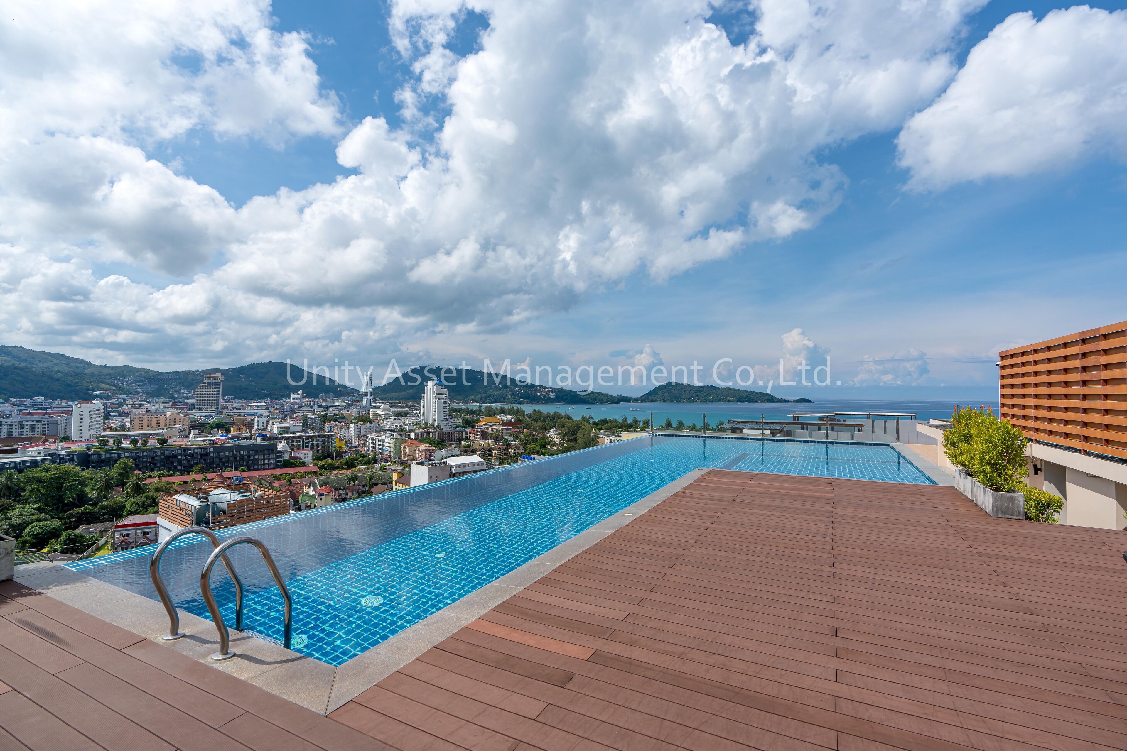 A rooftop infinity pool with a wooden deck, overlooking a cityscape and a body of water.