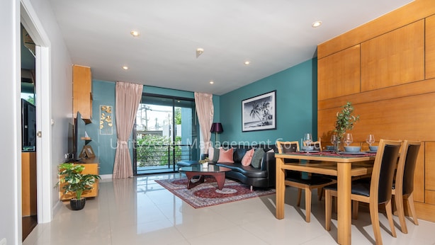 A modern living room with a dining area, teal walls, wooden paneling, a large window, and a view of greenery. - The Unity & The Bliss Patong Residence (Patong)