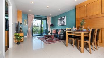 A modern living room with a dining area, teal walls, wooden paneling, a large window, and a view of greenery.