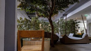 A modern indoor space with a wooden chair, a wicker sofa, and a large tree. - Brighton Hotel Bangkok (Bangkok)