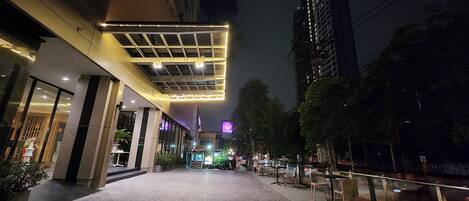 A nighttime urban street with a modern building featuring a glass facade and outdoor seating area.