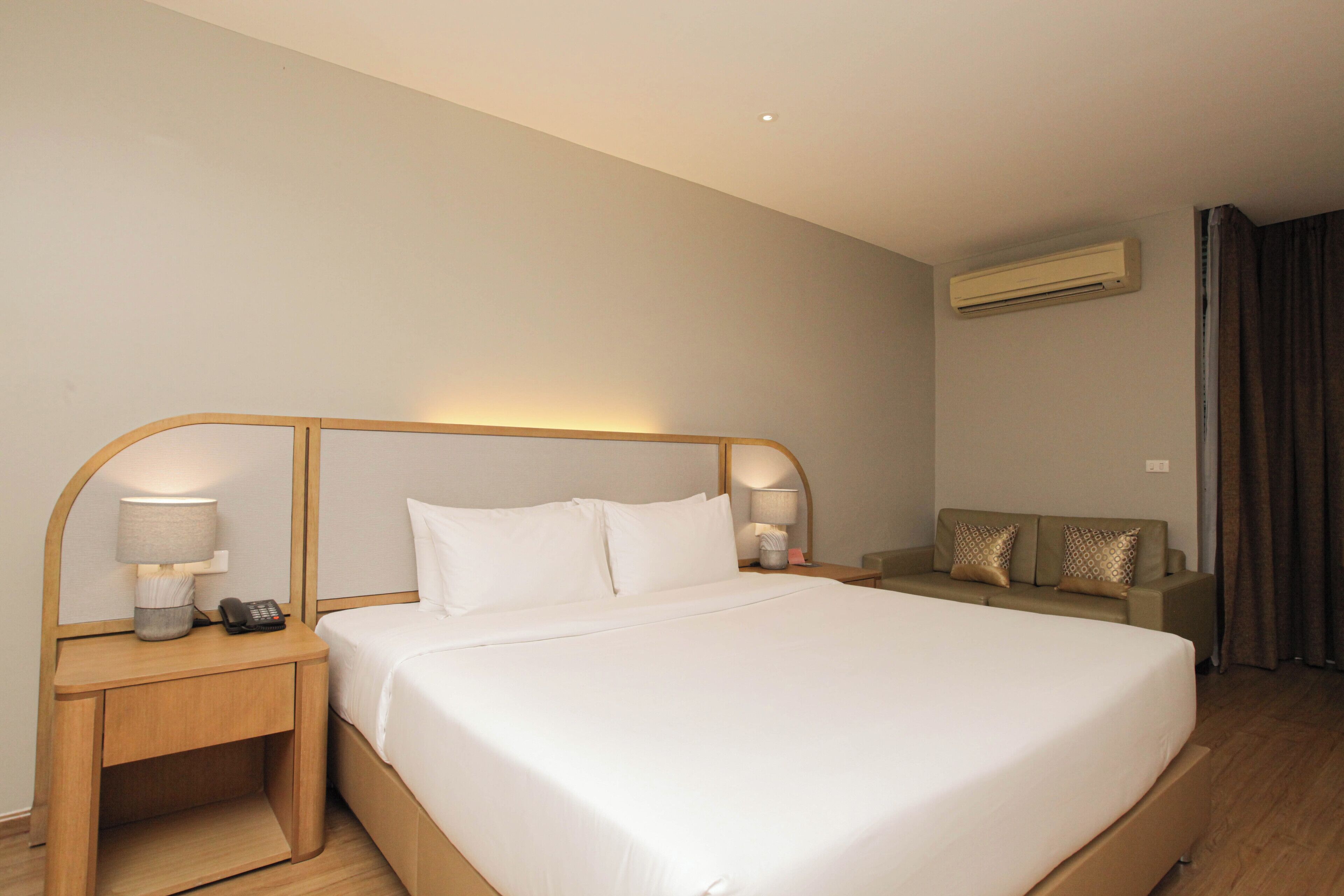 A hotel room with a large bed, two bedside tables with lamps, a sofa, and an air conditioning unit.