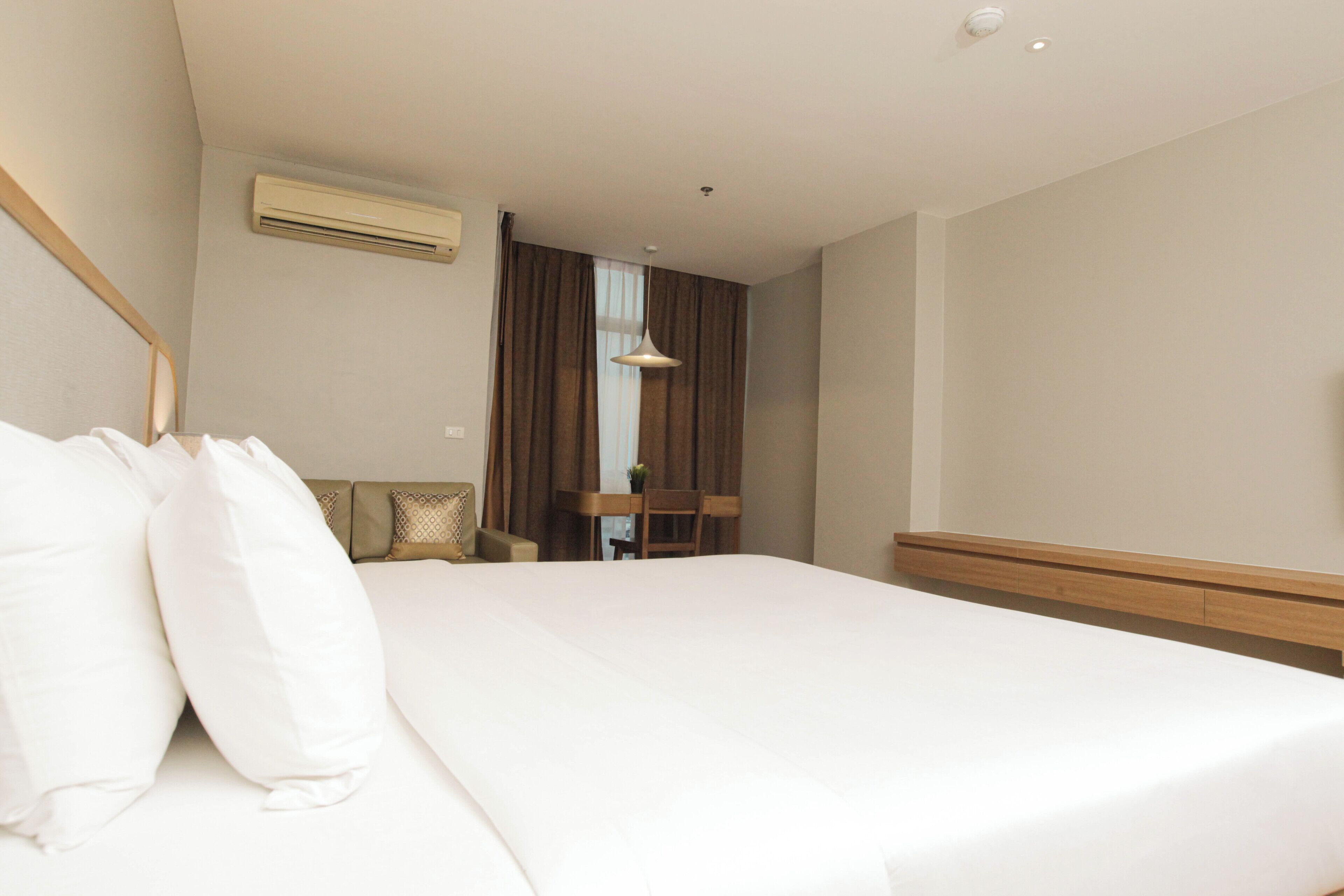 A hotel room with a large bed, a desk, and a chair. There is an air conditioning unit on the wall and a hanging light fixture.