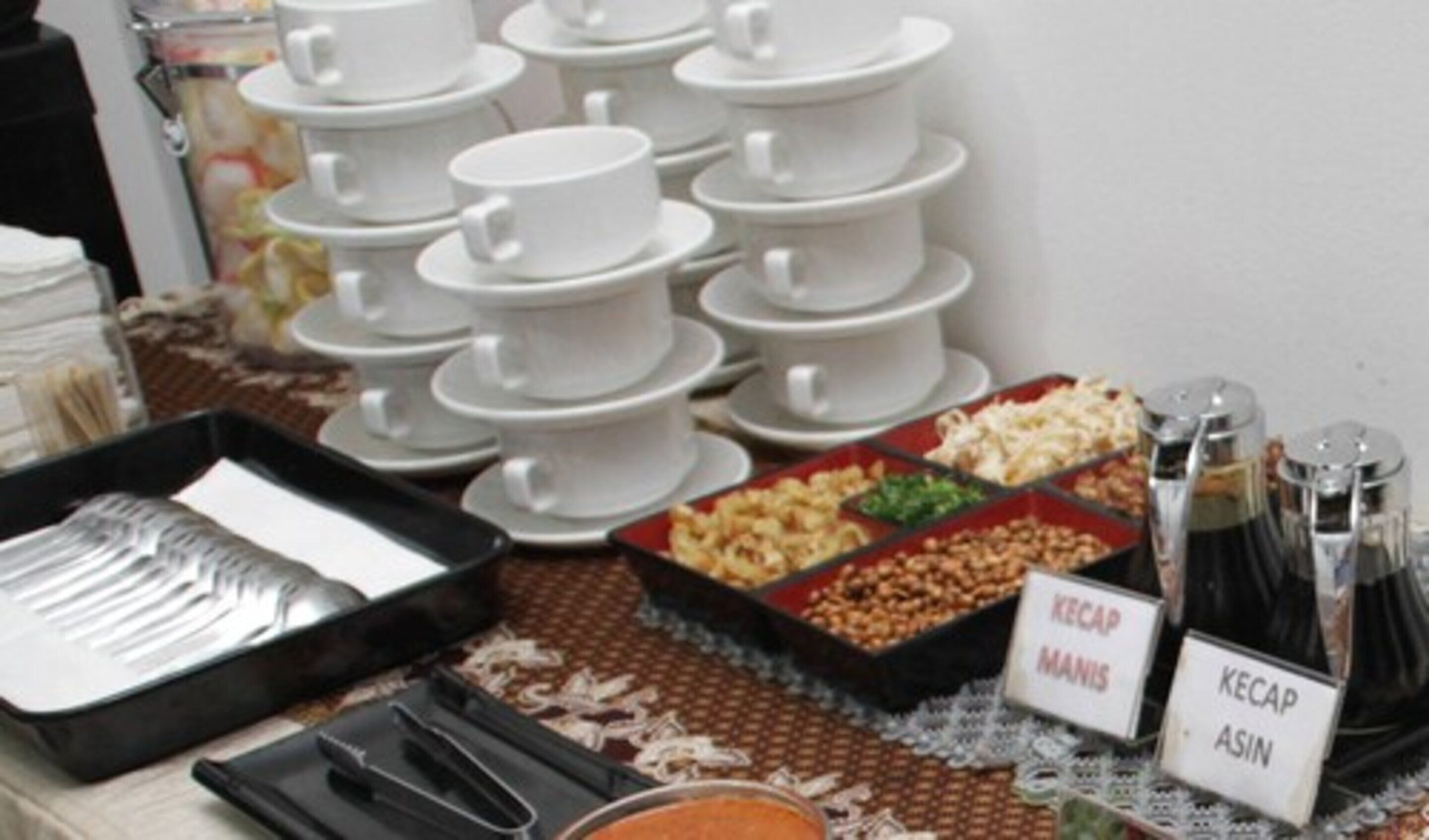 A buffet setup with multiple white bowls and plates, a tray of utensils, and labeled containers of Kecap Manis and Kecap Asin.