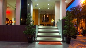 Entrada de un hotel con una escalera que lleva al vestíbulo, plantas en macetas a ambos lados y una alfombra roja al final.