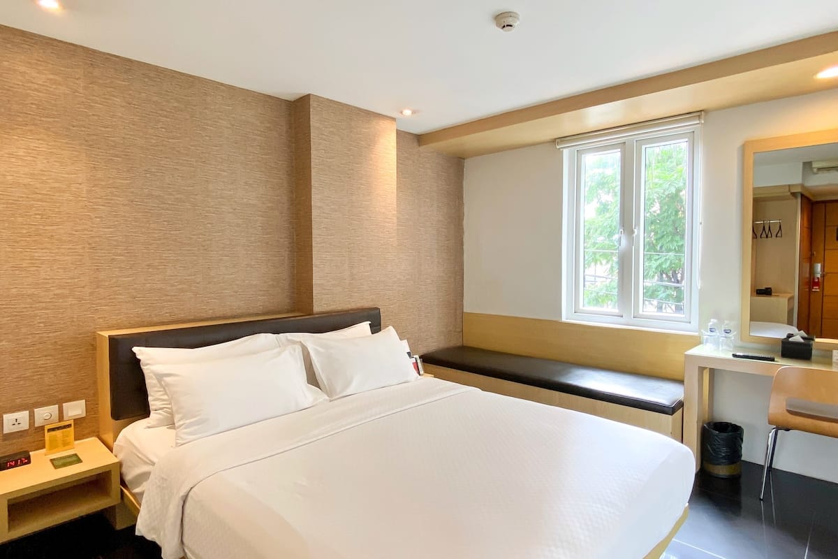 A modern hotel room with a large bed, a desk, and a window with a view of greenery.