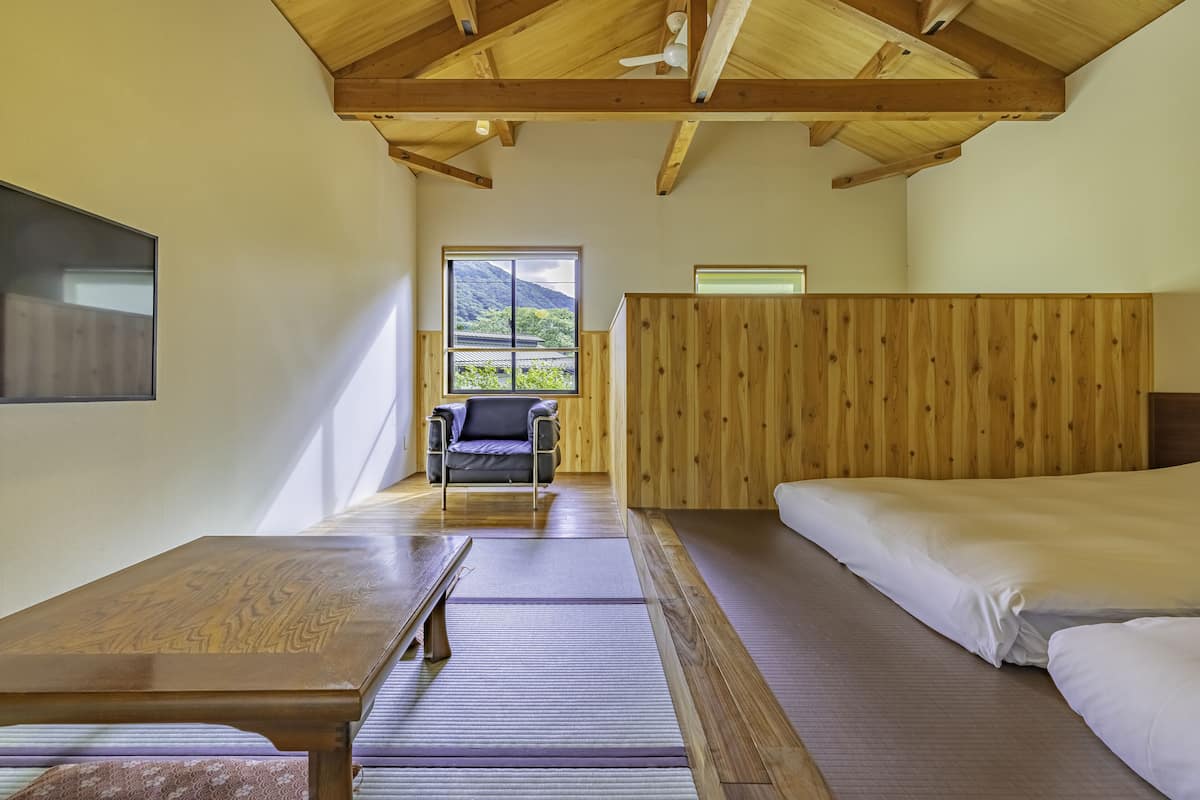 A room with a wooden ceiling, a bed, a chair, a table, and a window with a view of greenery.