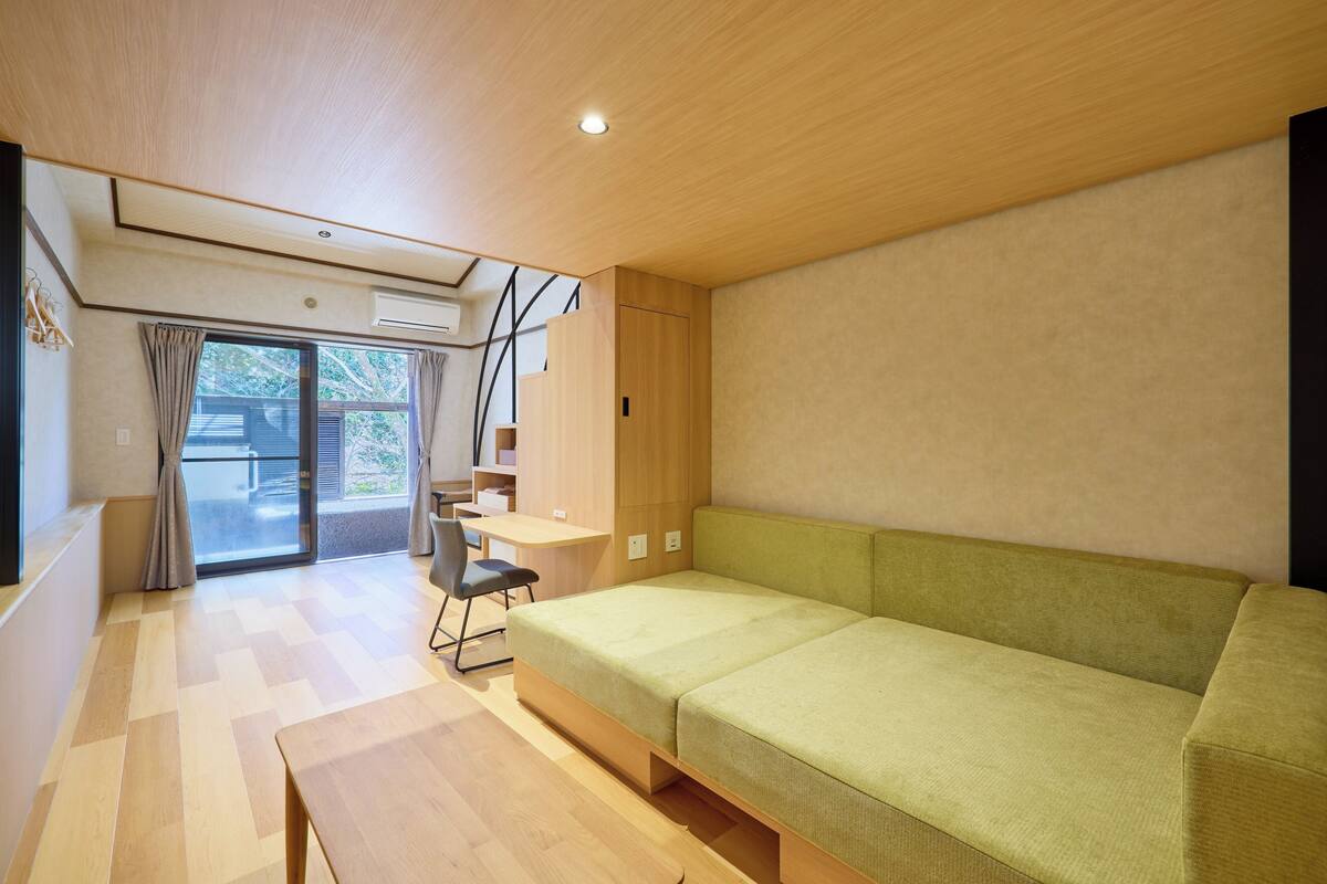 A modern room with a green sofa, wooden flooring, a desk, and a sliding glass door leading to an outdoor area.