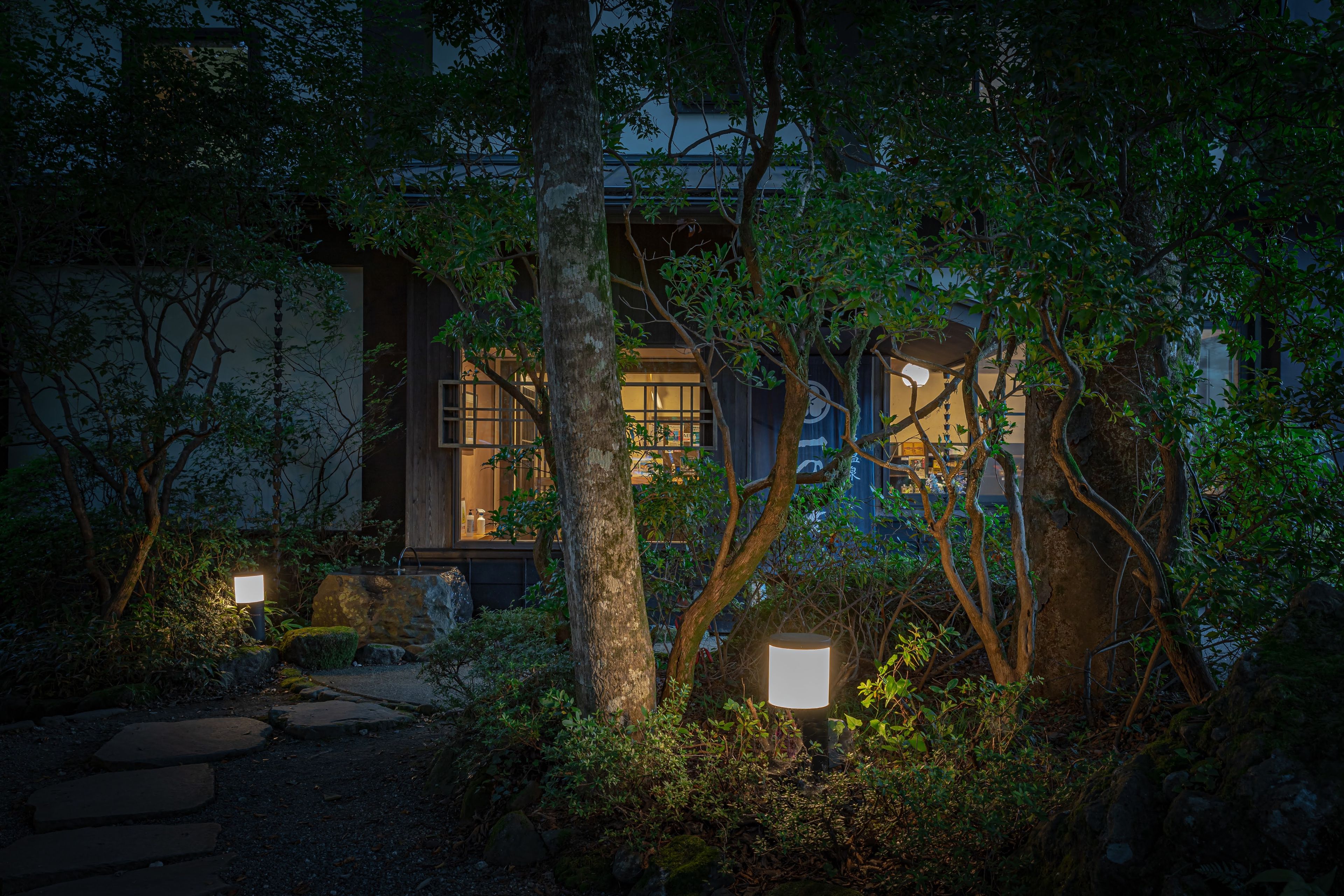 A traditional Japanese-style building with a stone pathway leading to the entrance, surrounded by lush greenery and illuminated by soft lighting.