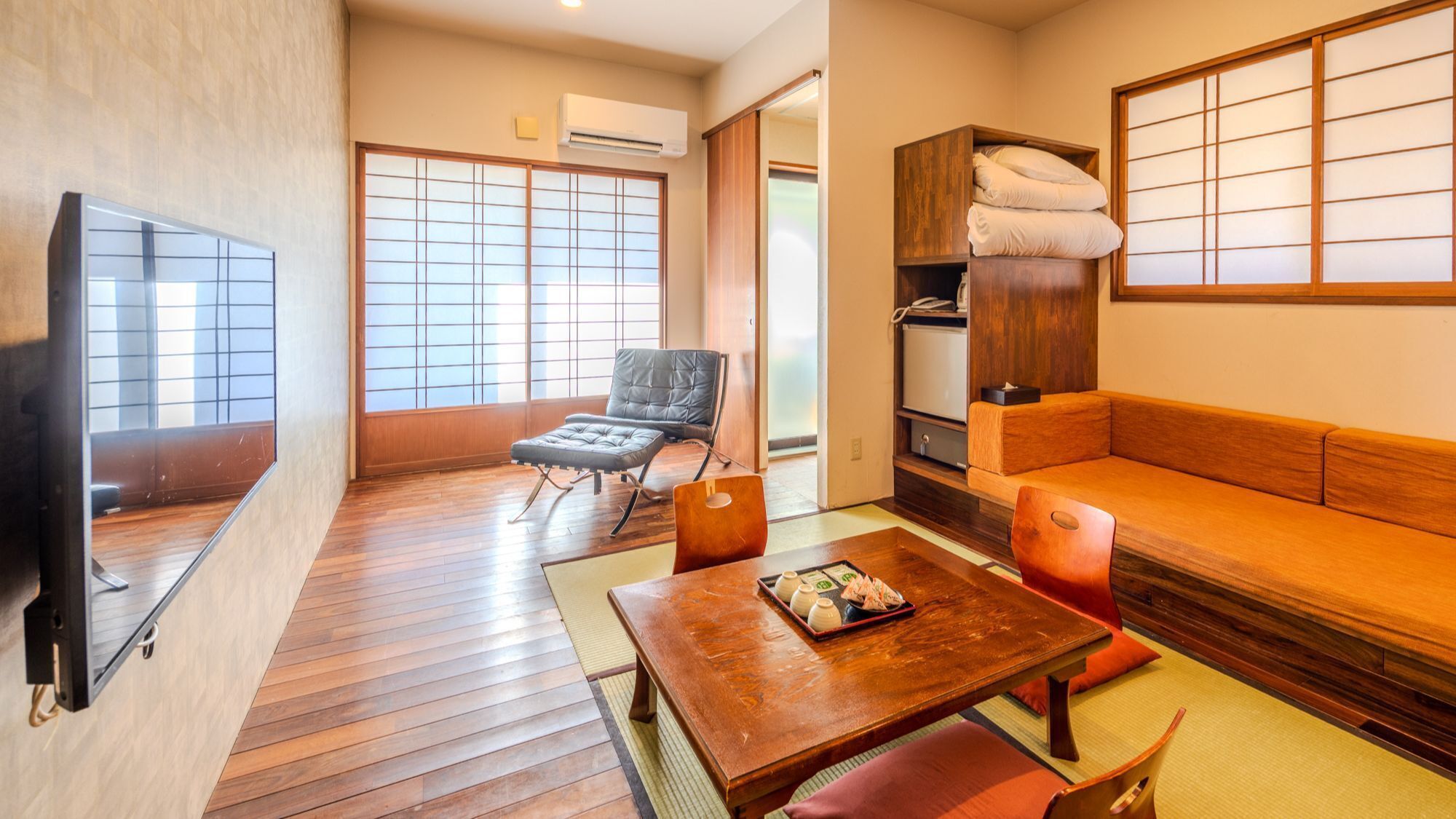 A traditional Japanese-style room with tatami flooring, a low table with a tray, a sofa, a chair, and a television.