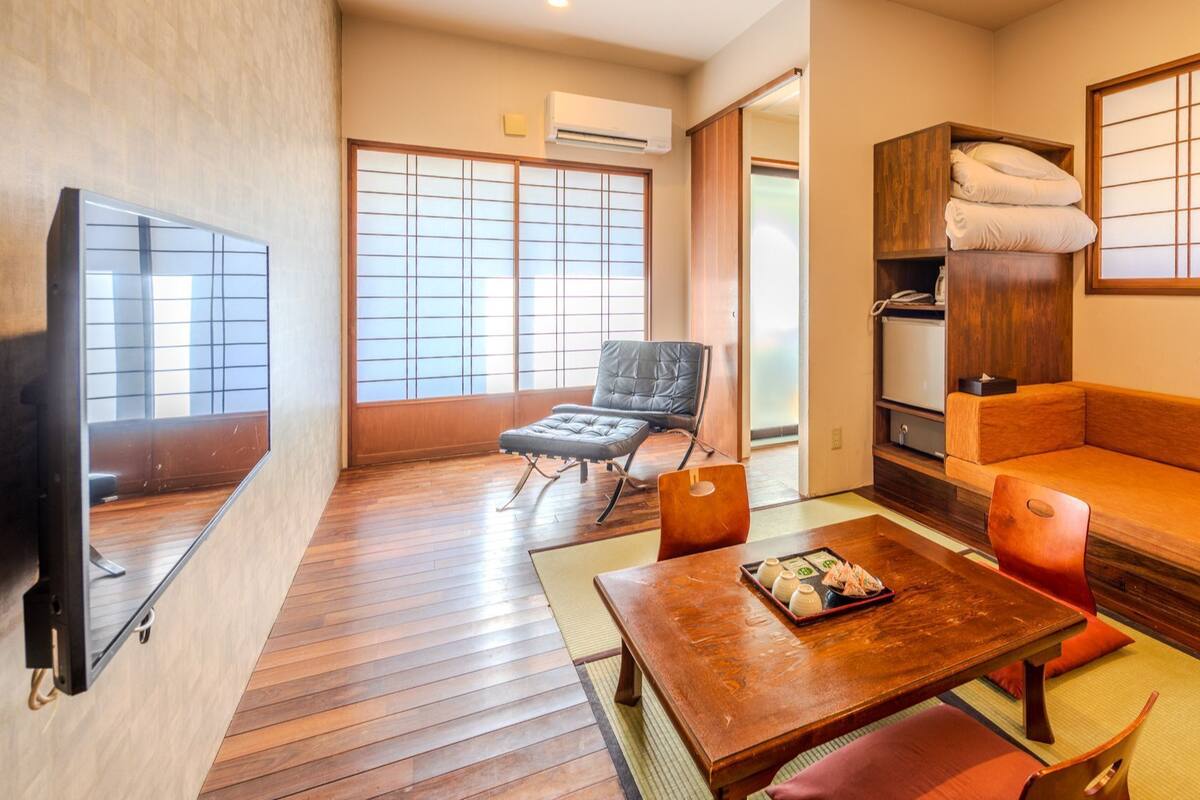 A traditional Japanese-style room with tatami flooring, a low table with a tray, a sofa, a chair, and a television.