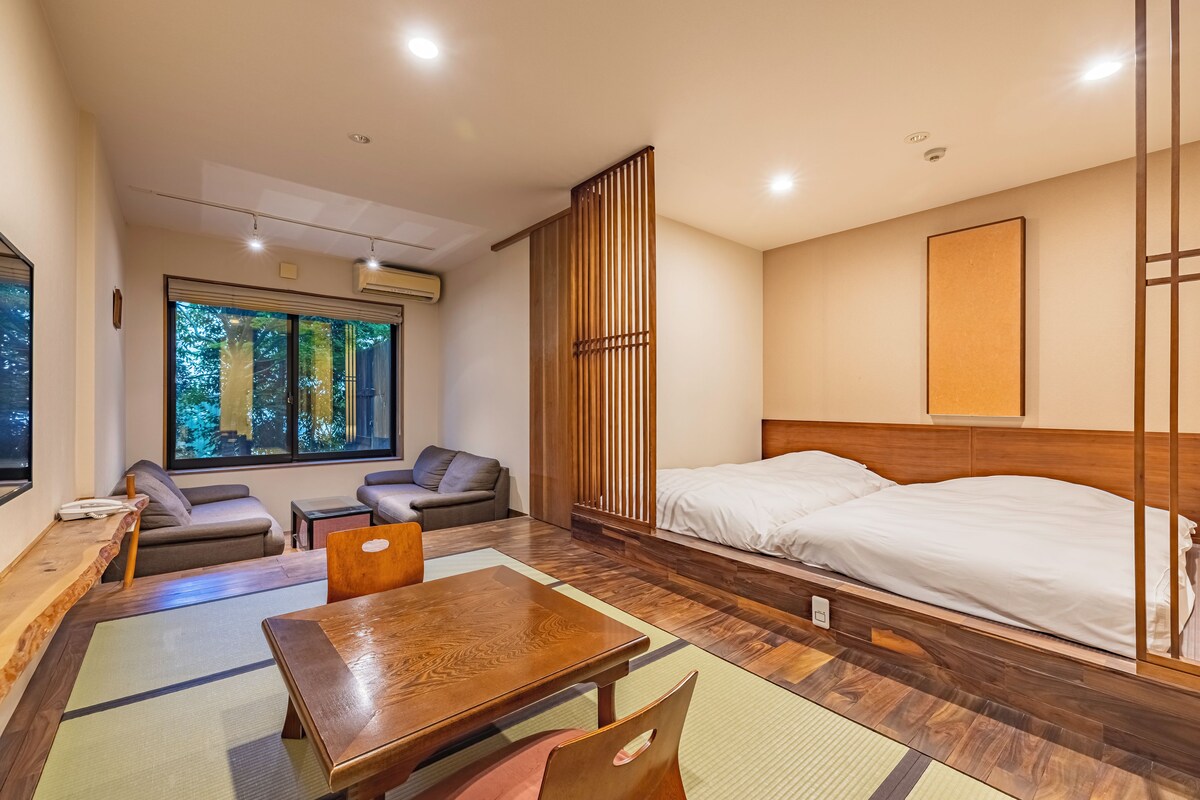 Quarto de hotel moderno com uma cama grande, uma mesa de madeira, um sofá e vista para árvores pela janela.