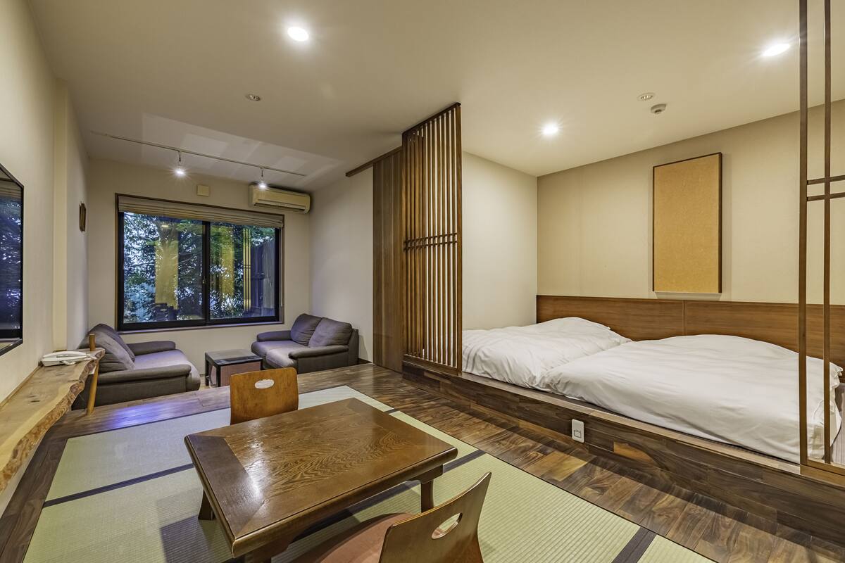A modern hotel room with a large bed, a wooden table, a sofa, and a view of trees through the window.