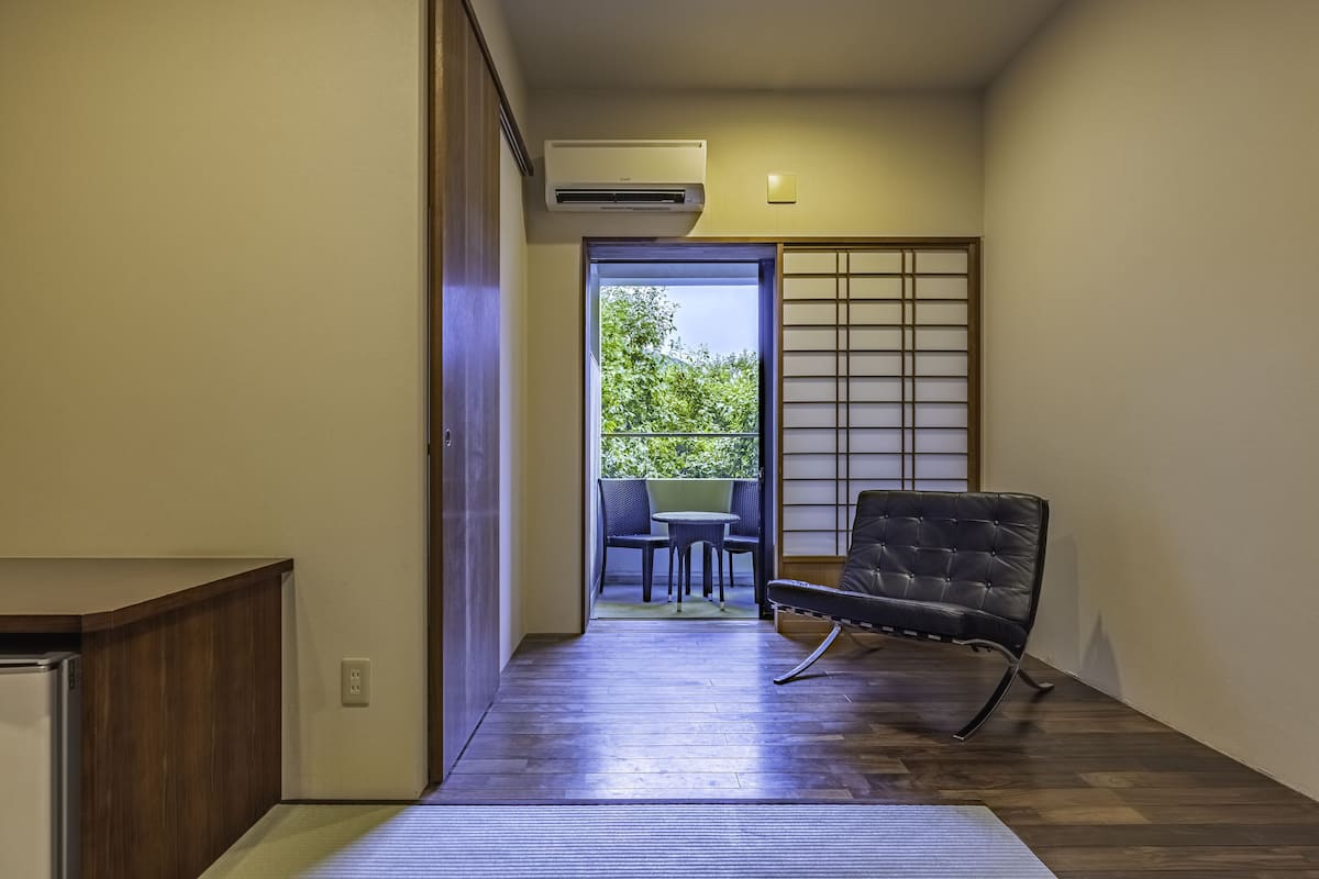 A hallway with a wooden floor, a black chair, a small table, and a sliding door leading to an outdoor area with a view of trees.