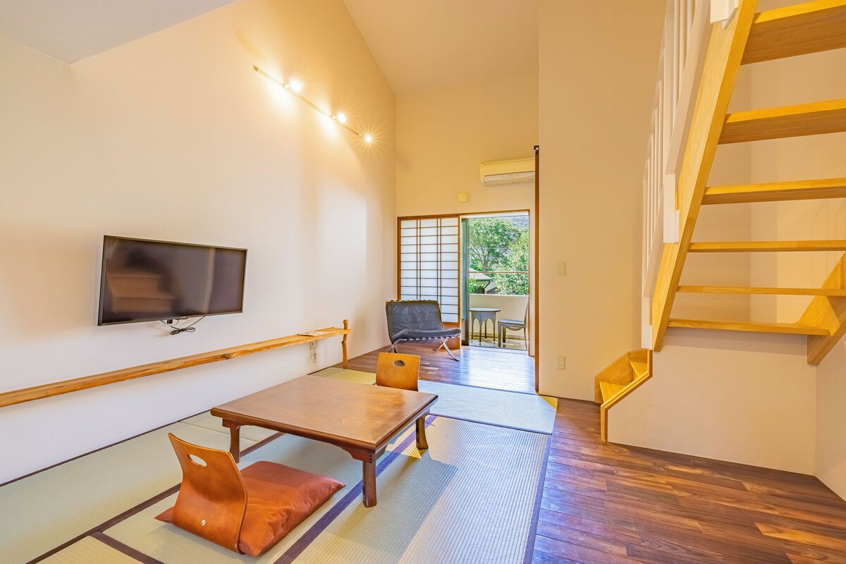 Um quarto amplo com uma mesa de madeira, um sofá, uma TV fixada na parede e uma escada.