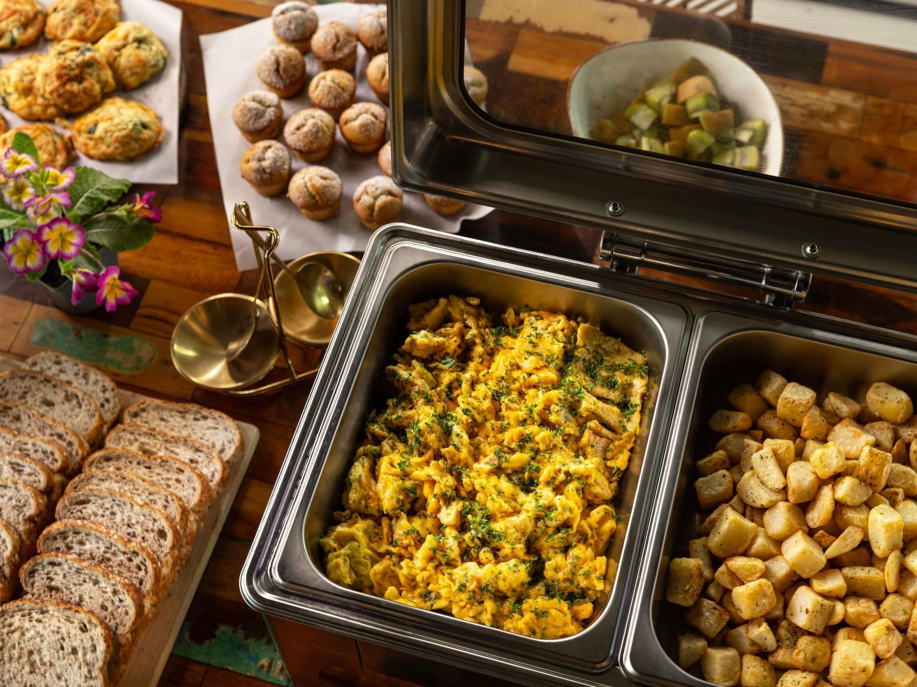 A buffet table with various food items including scrambled eggs, roasted potatoes, and bread.