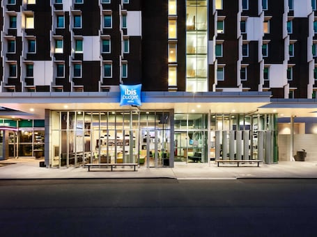 A modern hotel with a glass facade, a blue sign indicating it's an ibis budget hotel, and a well-lit entrance.. ibis budget Auckland Airport