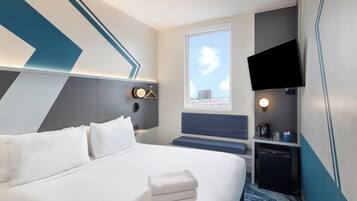 A modern hotel room with a bed, a desk, a television, and a window with a city view.