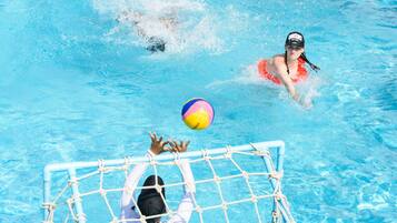 Una piscina con agua azul cristalina, una portería y una persona con camisa blanca alcanzando una pelota.