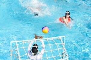 Una piscina con agua azul cristalina, una portería y una persona con camisa blanca alcanzando una pelota.