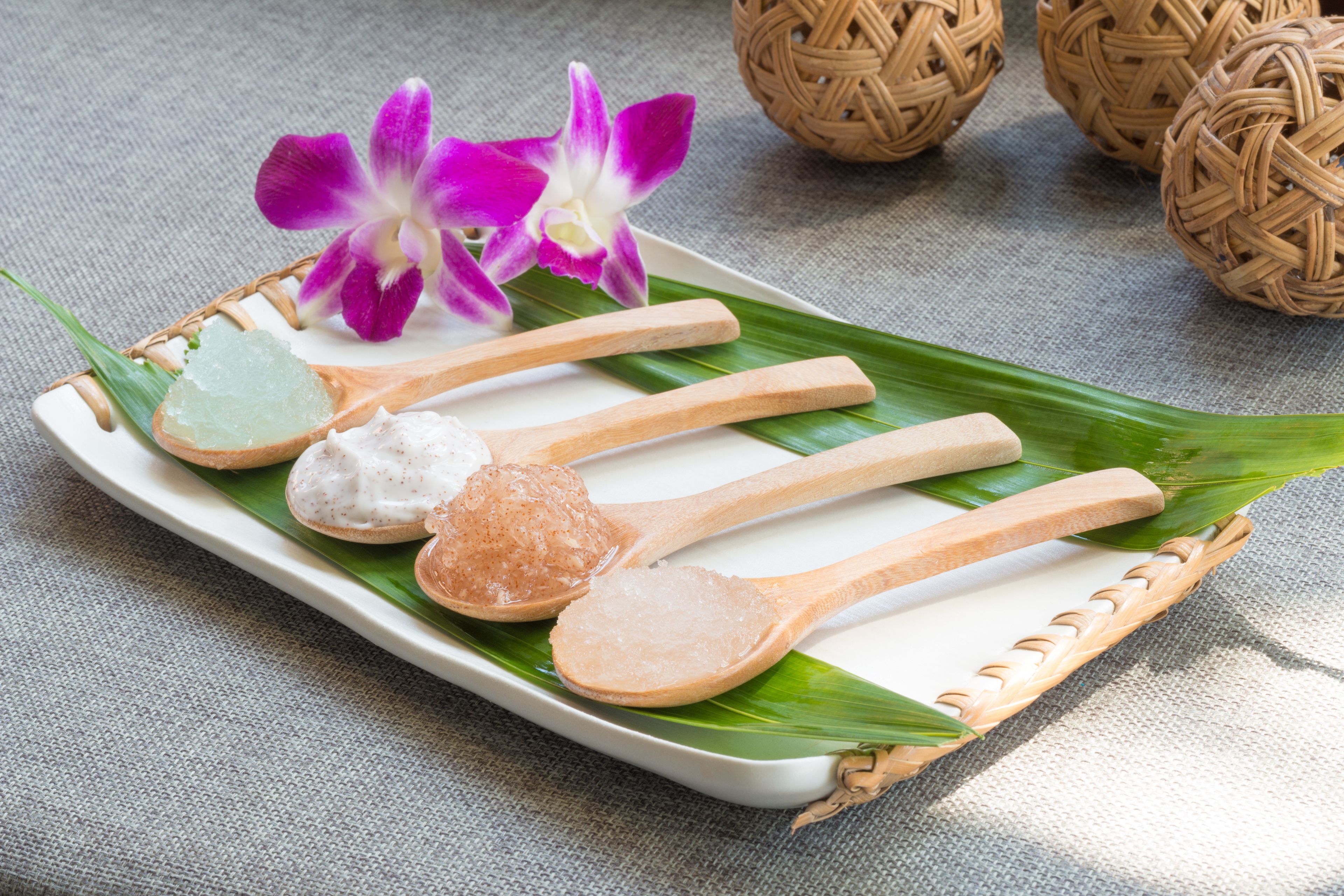 A tray with wooden spoons and a white plate on a green leaf, with a purple orchid and woven balls in the background.