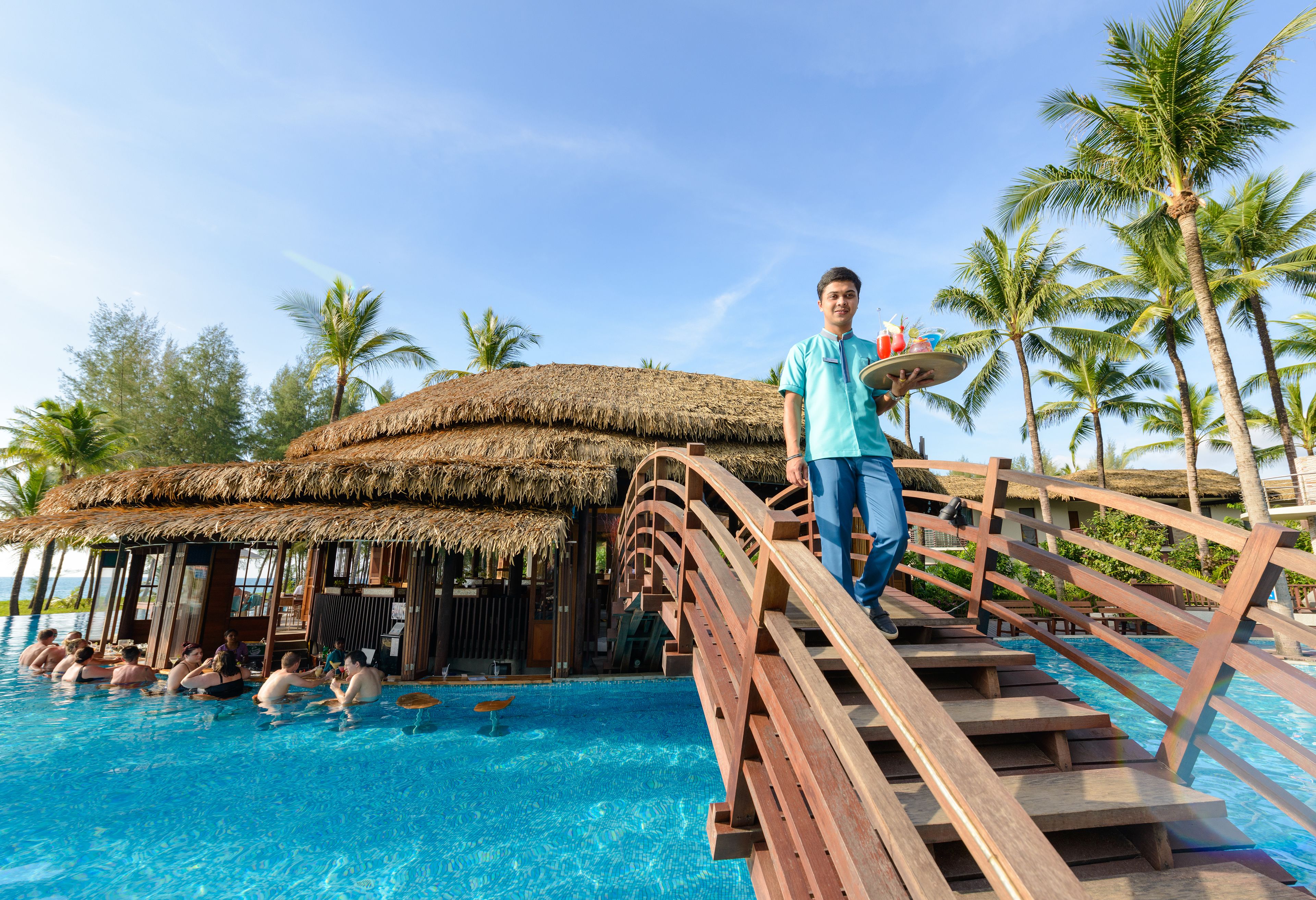 A wooden bridge over a swimming pool with palm trees and a thatched-roof structure in the background.