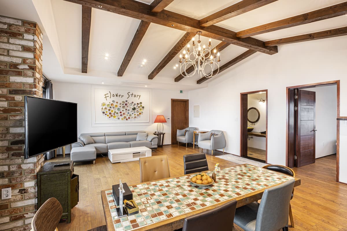 A spacious living room with a large flat-screen TV, a sofa, a dining table with chairs, and a chandelier.