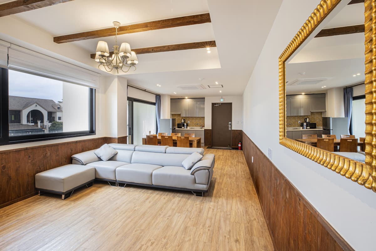 A modern living room with a sectional sofa, wooden flooring, a large window, and a kitchen visible through an open door.