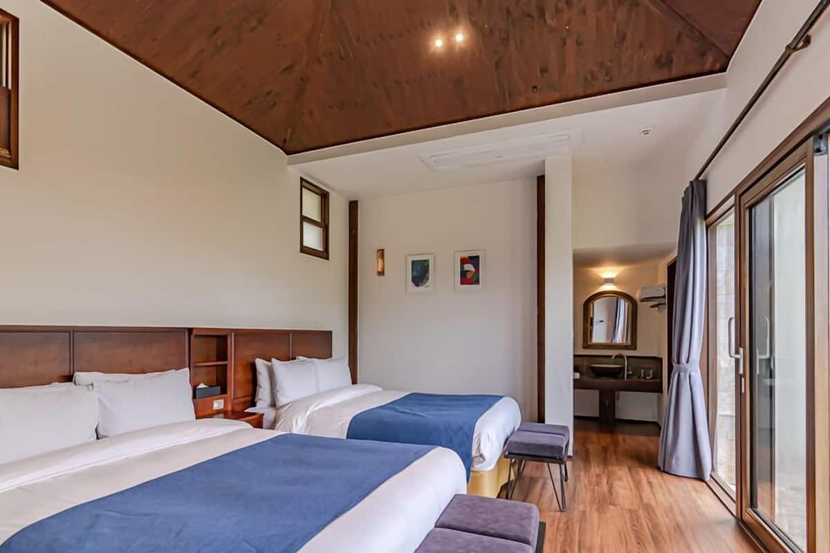 A hotel room with two beds, a wooden ceiling, a bathroom with a sink and mirror, and artwork on the walls.