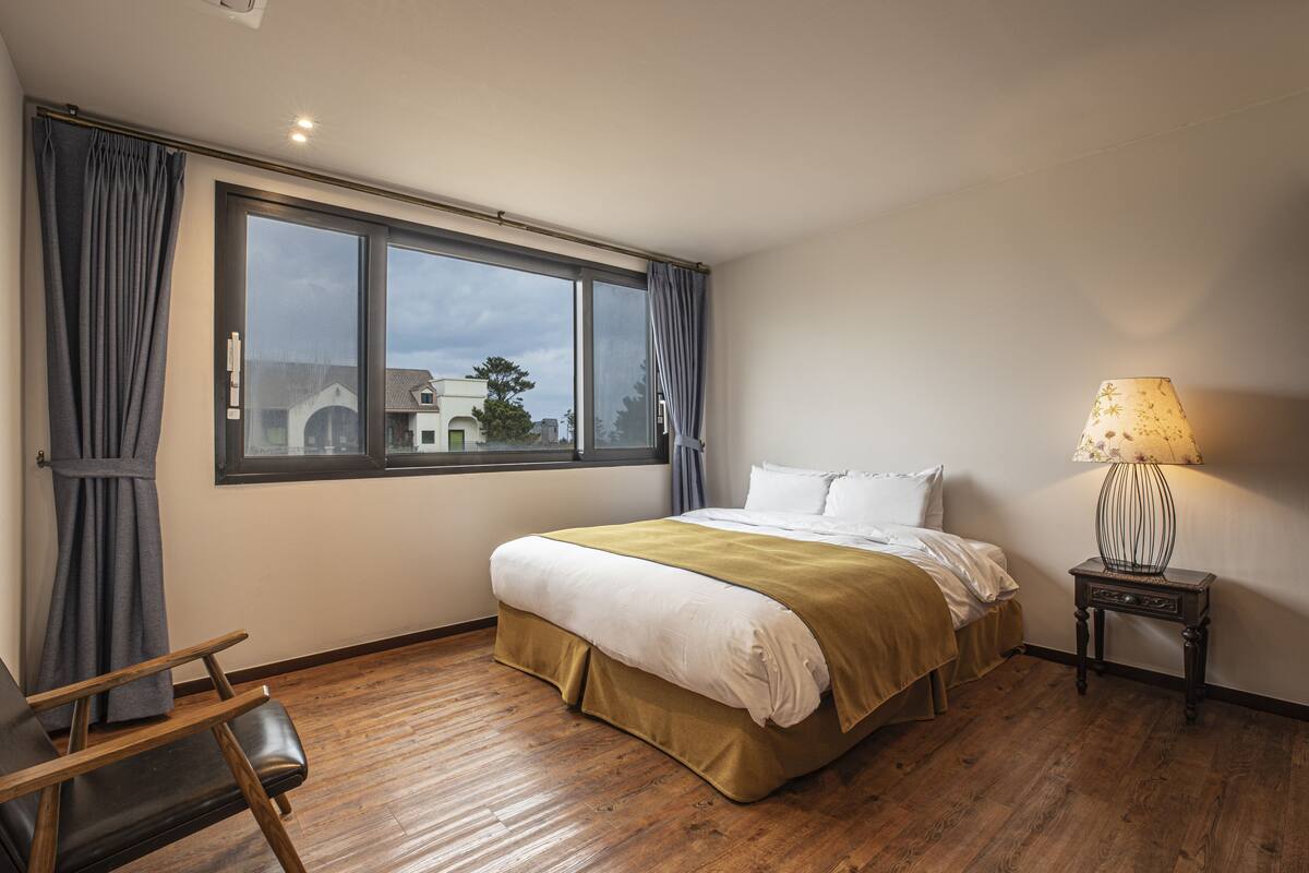 A bedroom with a large window, a bed, a chair, and a nightstand with a lamp.