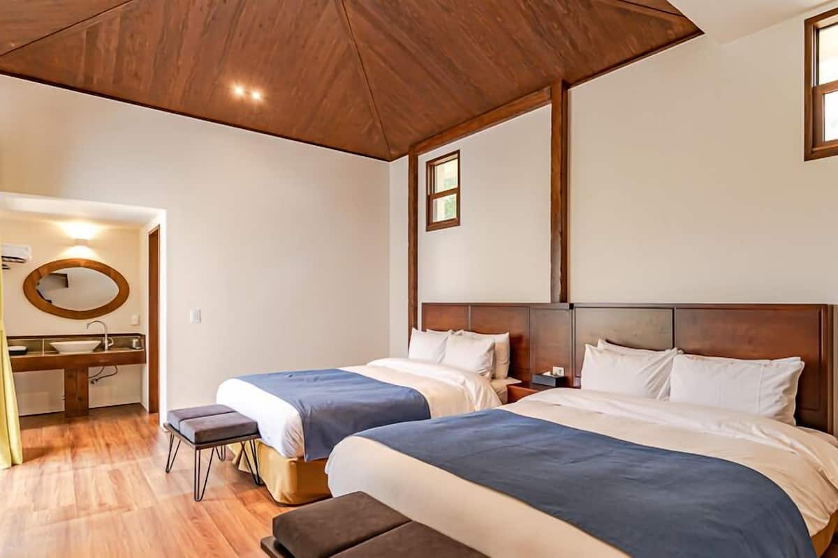 A hotel room with two beds, a wooden ceiling, a bathroom with a mirror and sink, and a window.