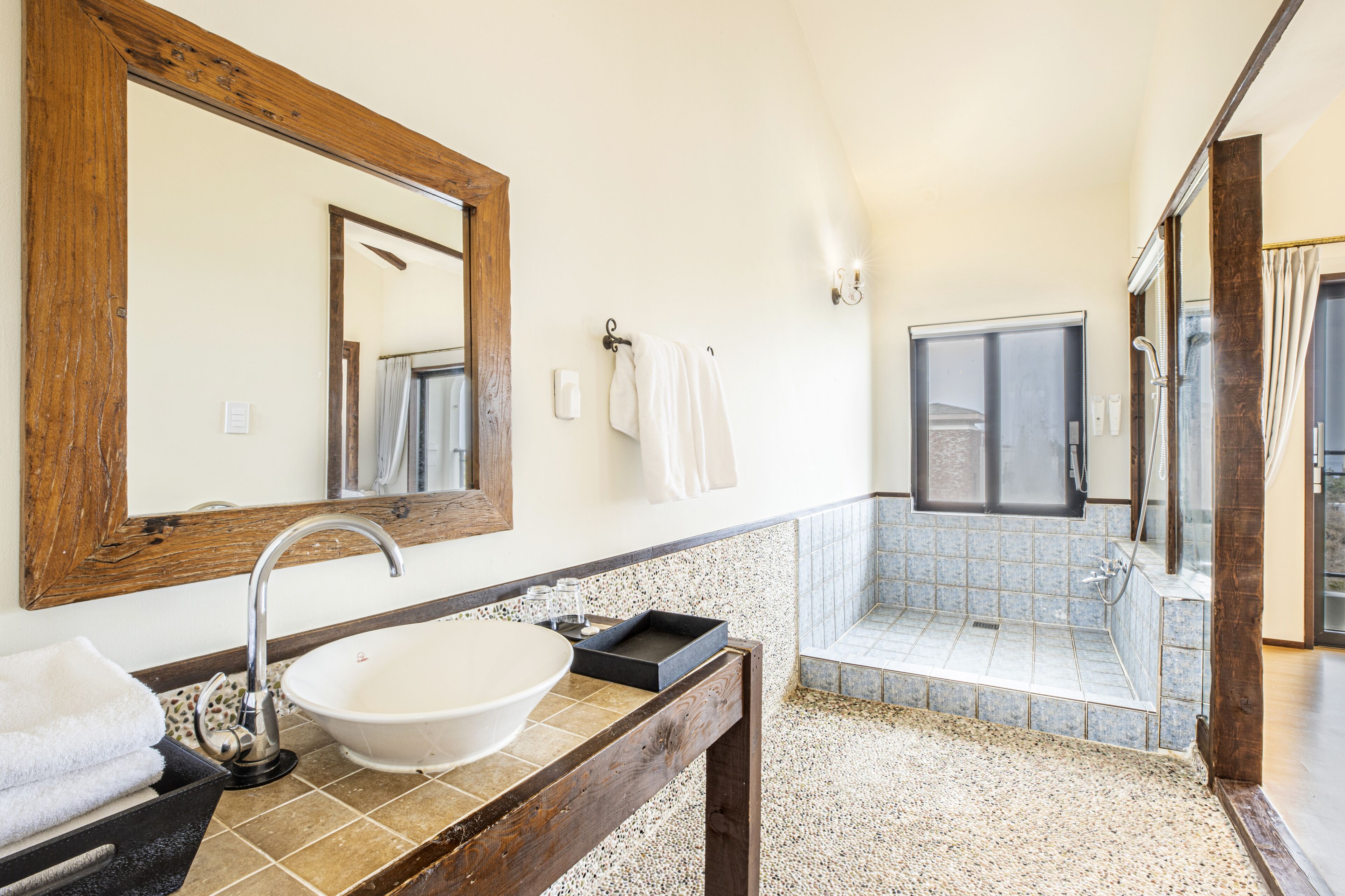 A bathroom with a wooden vanity, a white sink, and a large mirror. There is a shower area with a glass door and a tiled floor.