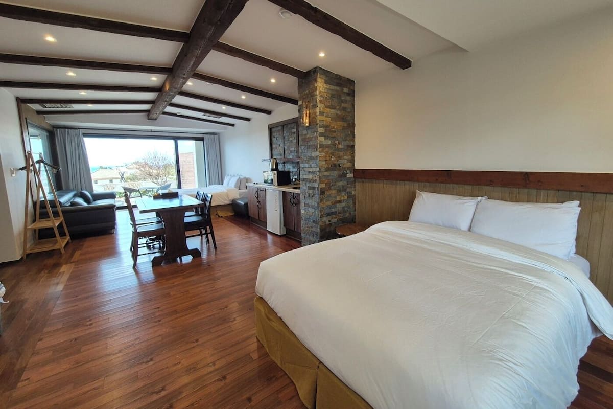 A modern hotel room with a large bed, wooden flooring, a stone accent wall, a dining area with a table and chairs, and a view through a large window.