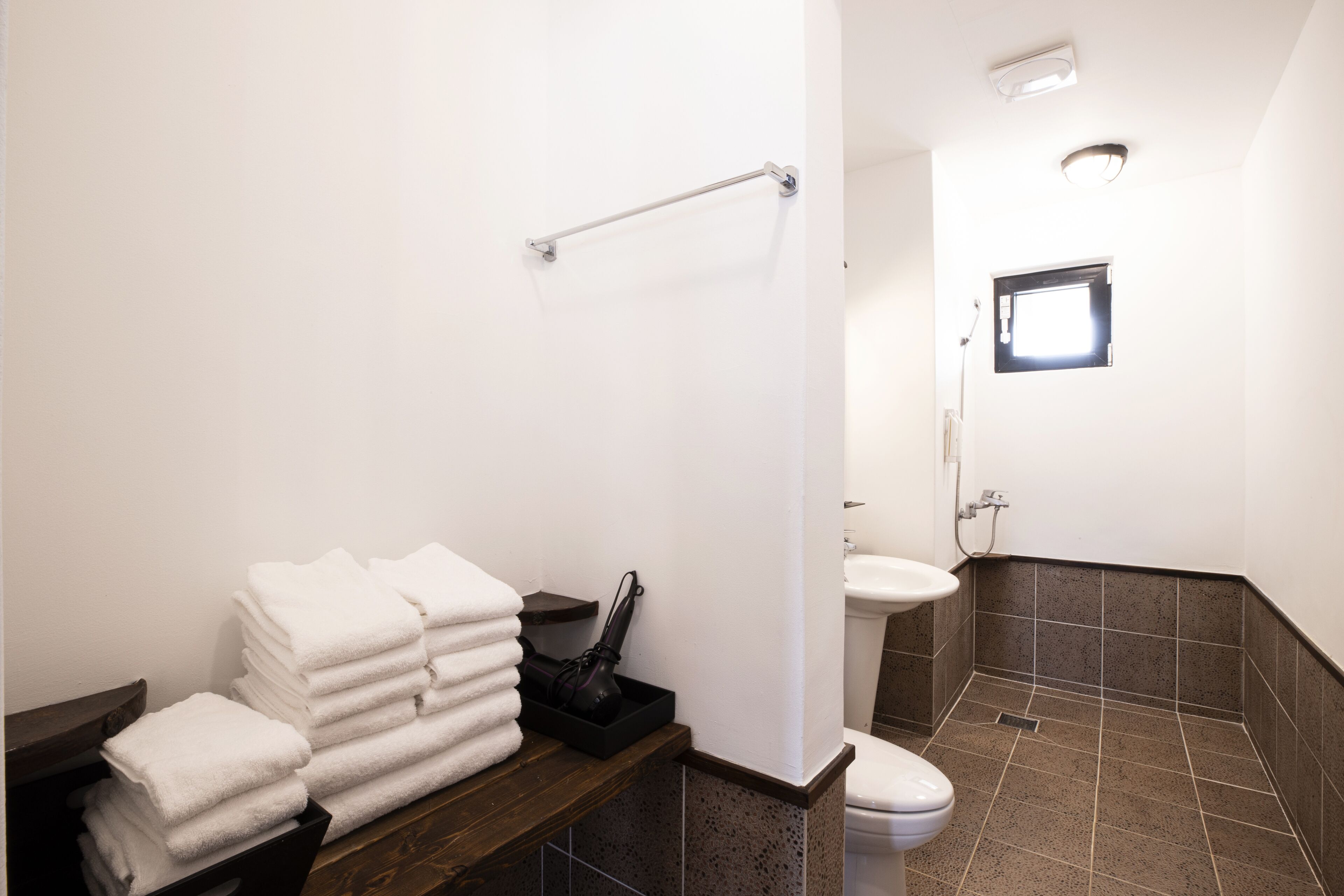 A bathroom with a toilet, sink, and a small window. There is a towel rack with folded towels and a black container.