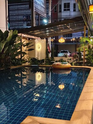 A swimming pool with a tiled deck, surrounded by plants and a covered area with string lights. - Chiang Mai Waroros Boutique Hotel (Chiang Mai)