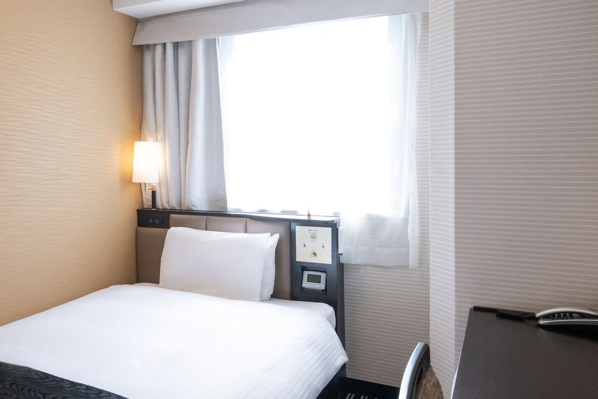 Une chambre d’hôtel avec un lit, un bureau, une lampe et une fenêtre avec des rideaux.