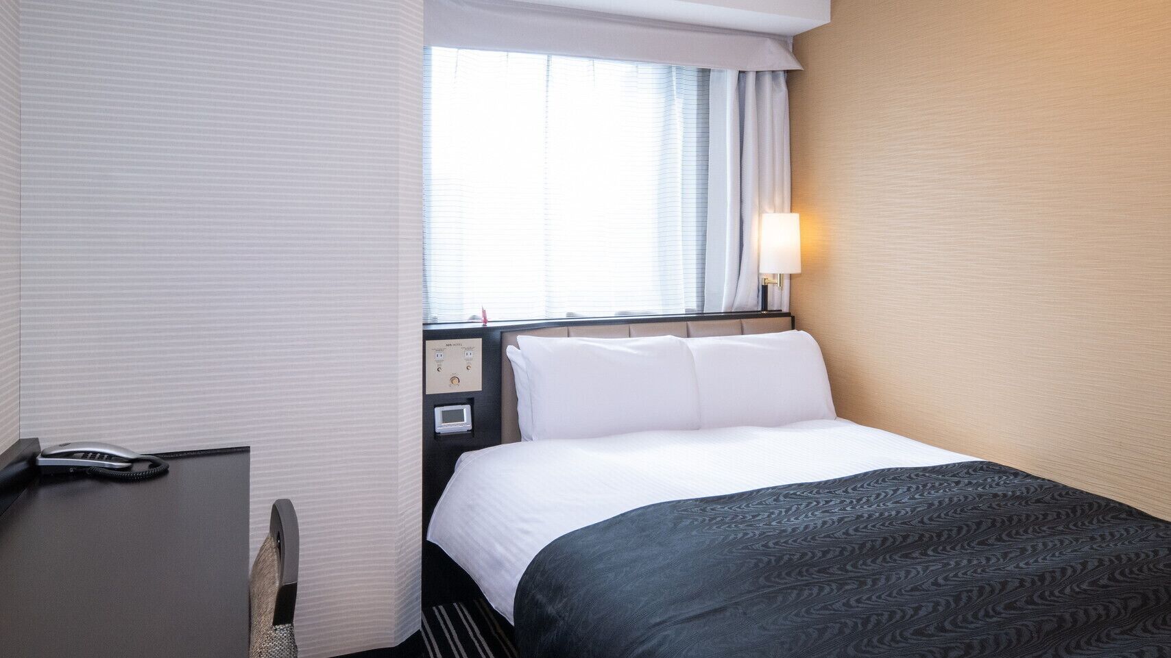 Une chambre d’hôtel comprenant un lit, une table de chevet, une lampe, une fenêtre avec des rideaux et un panneau de commande au mur.