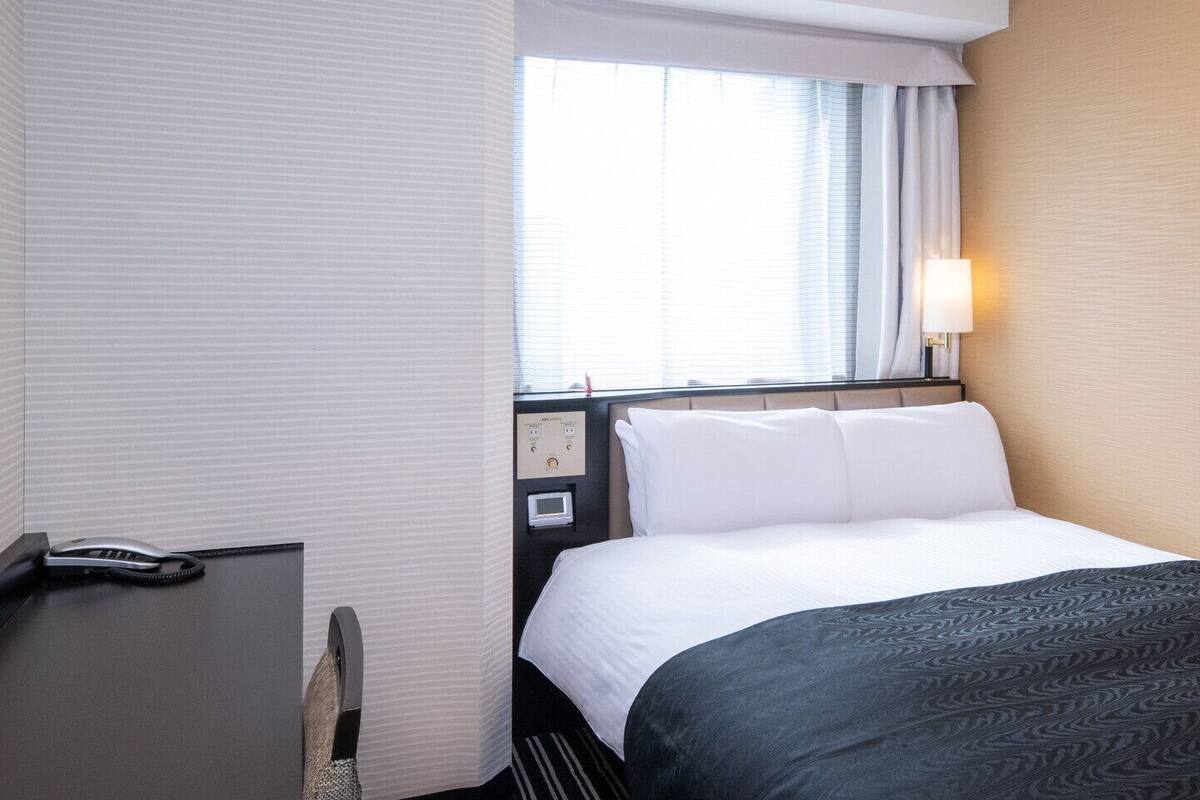 Une chambre d’hôtel comprenant un lit, une table de chevet, une lampe, une fenêtre avec des rideaux et un panneau de commande au mur.