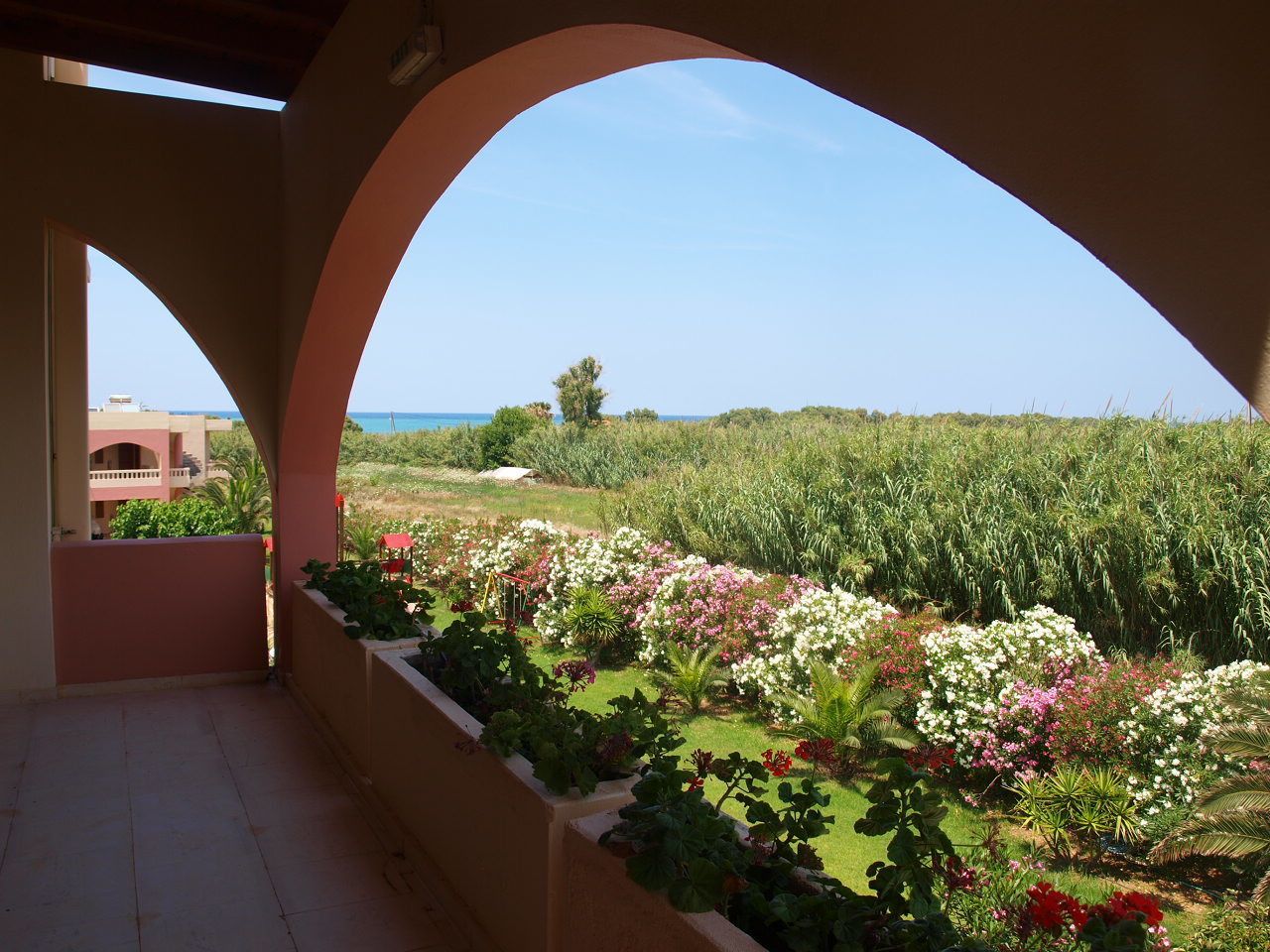 A covered balcony with a view of a garden and the sea.