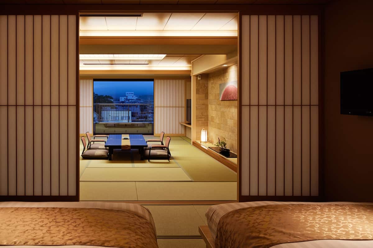 Une pièce de style japonais traditionnel, avec un sol recouvert de tatamis, une table basse entourée de chaises et une vue sur le paysage urbain.