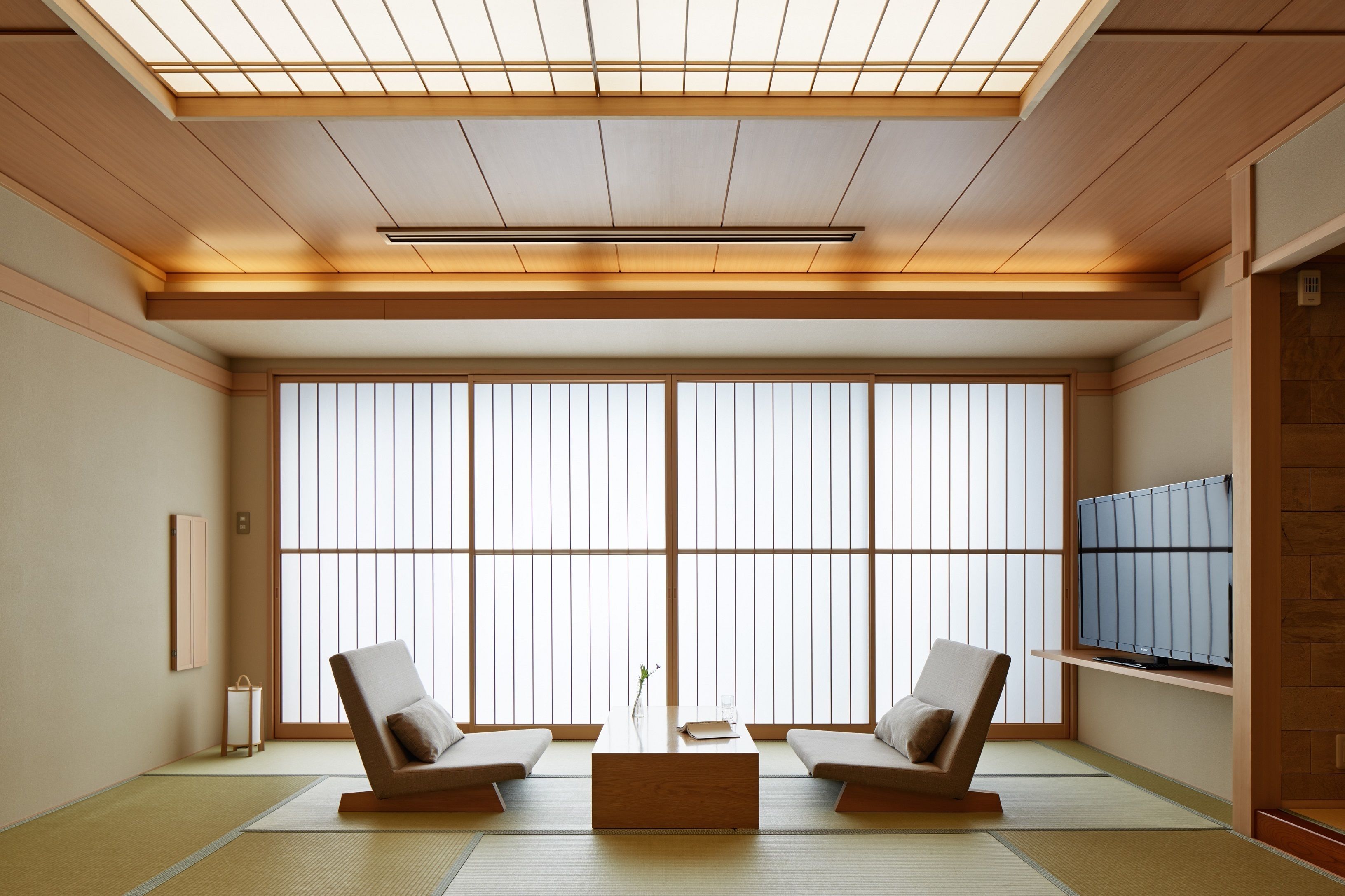 A traditional Japanese room with tatami flooring, a low wooden table, two cushioned chairs, and a TV on a shelf.