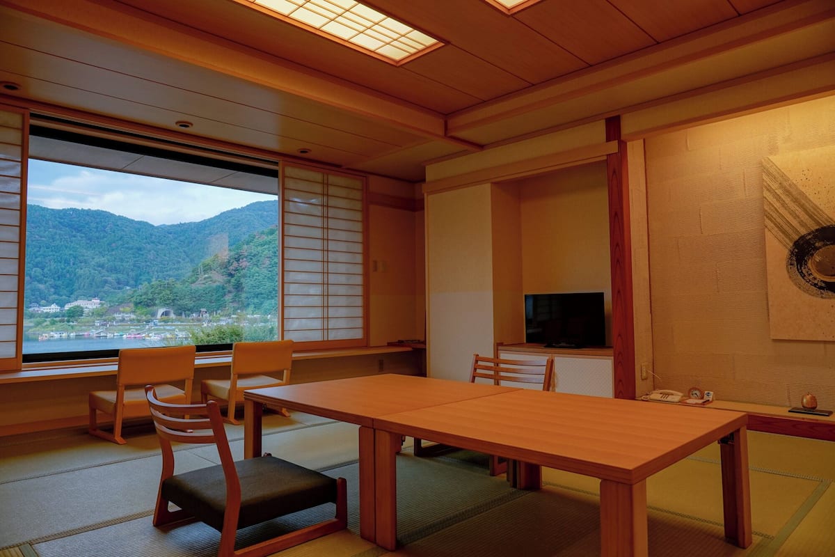 Une pièce de style japonais traditionnel, avec un sol recouvert de tatamis, une table basse et des chaises. Un téléviseur est fixé au mur, et une grande fenêtre offre une vue sur un paysage montagneux.