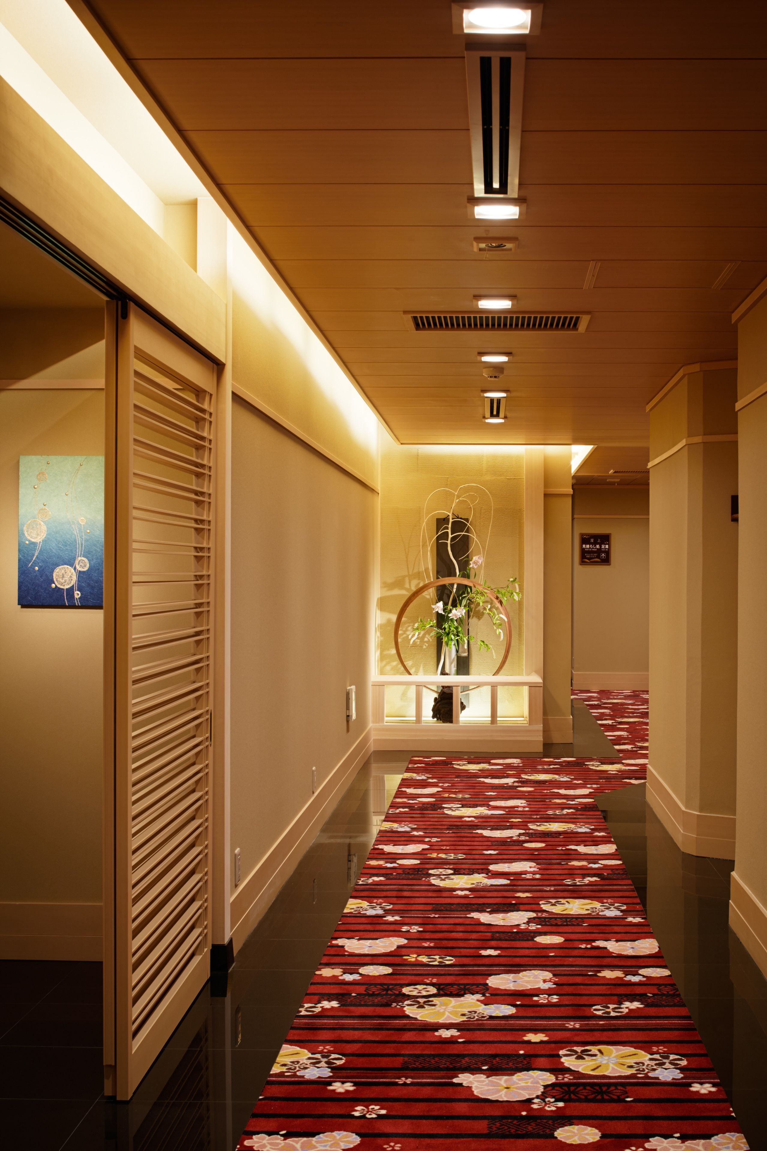 A hotel hallway with a red floral carpet, a wooden slatted door, and a wall-mounted artwork.