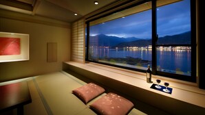 A room with a large window overlooking a body of water and mountains at dusk. There is a bed with a futon, pillows, and a bottle of wine on the windowsill. - Konansou (Fujikawaguchiko)