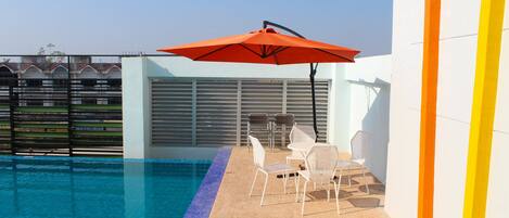 A swimming pool with a tiled edge, a seating area with chairs and a table, and a large orange umbrella.