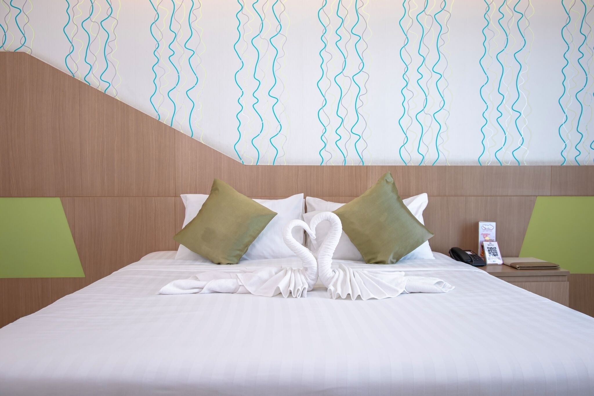 Una habitación de hotel con una cama bien hecha, almohadas verdes, un teléfono y una pequeña mesa con una libreta y un bolígrafo.