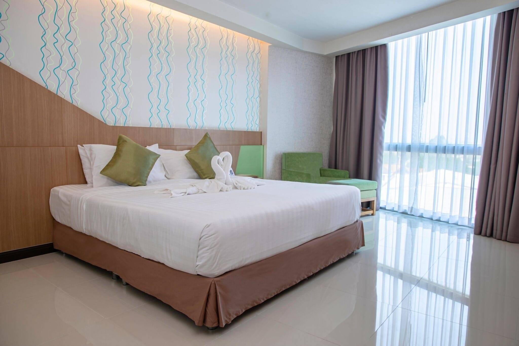 A hotel room with a large bed, a wooden headboard, a green armchair, and a view of the outdoors through sheer curtains.